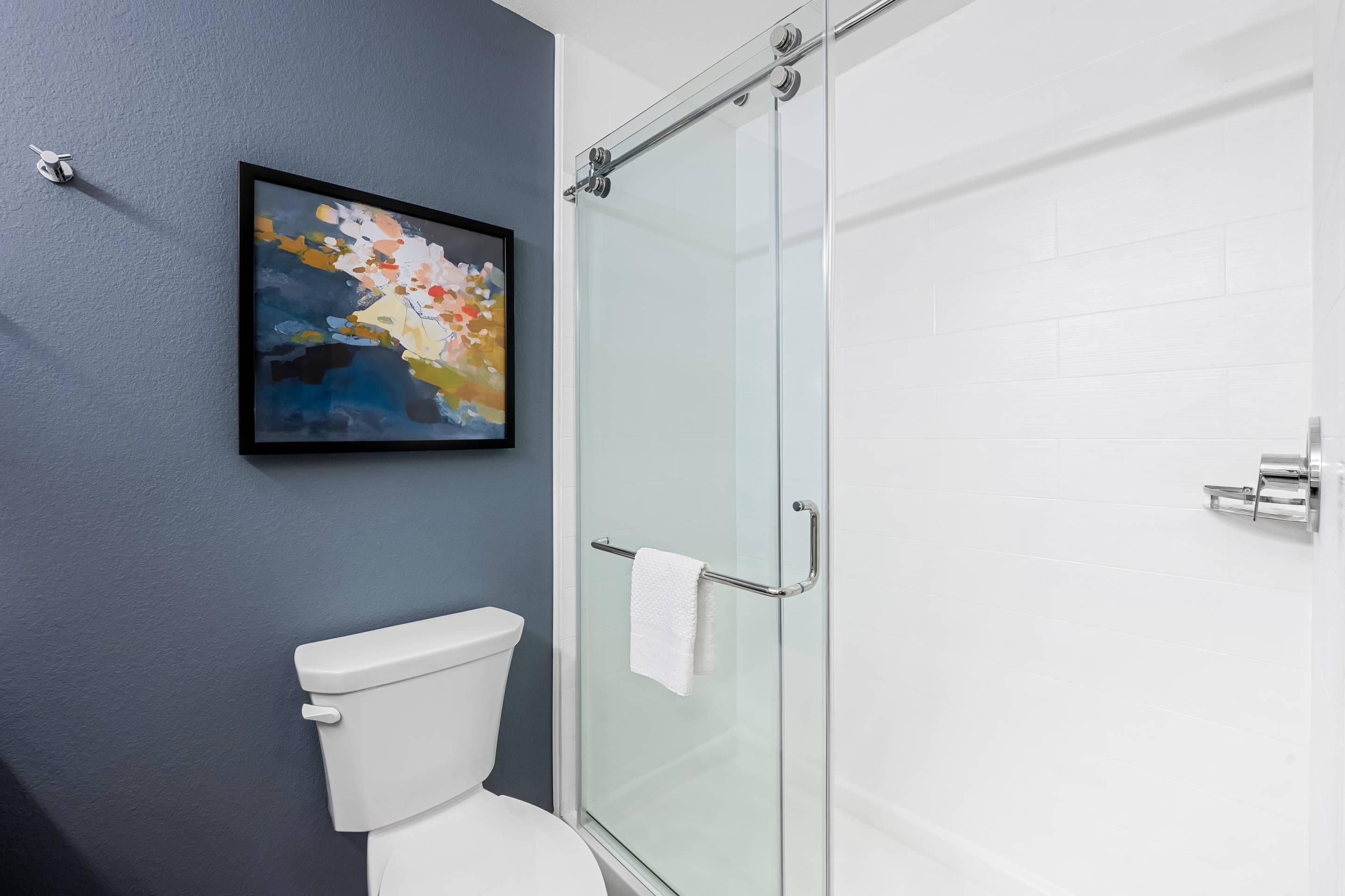 A bathroom with a glass shower enclosure, a white toilet, and a framed abstract painting on the wall.