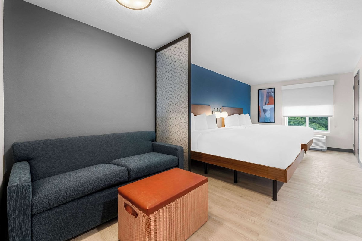 A modern hotel room with a grey sofa, a wooden bed, and a large window.