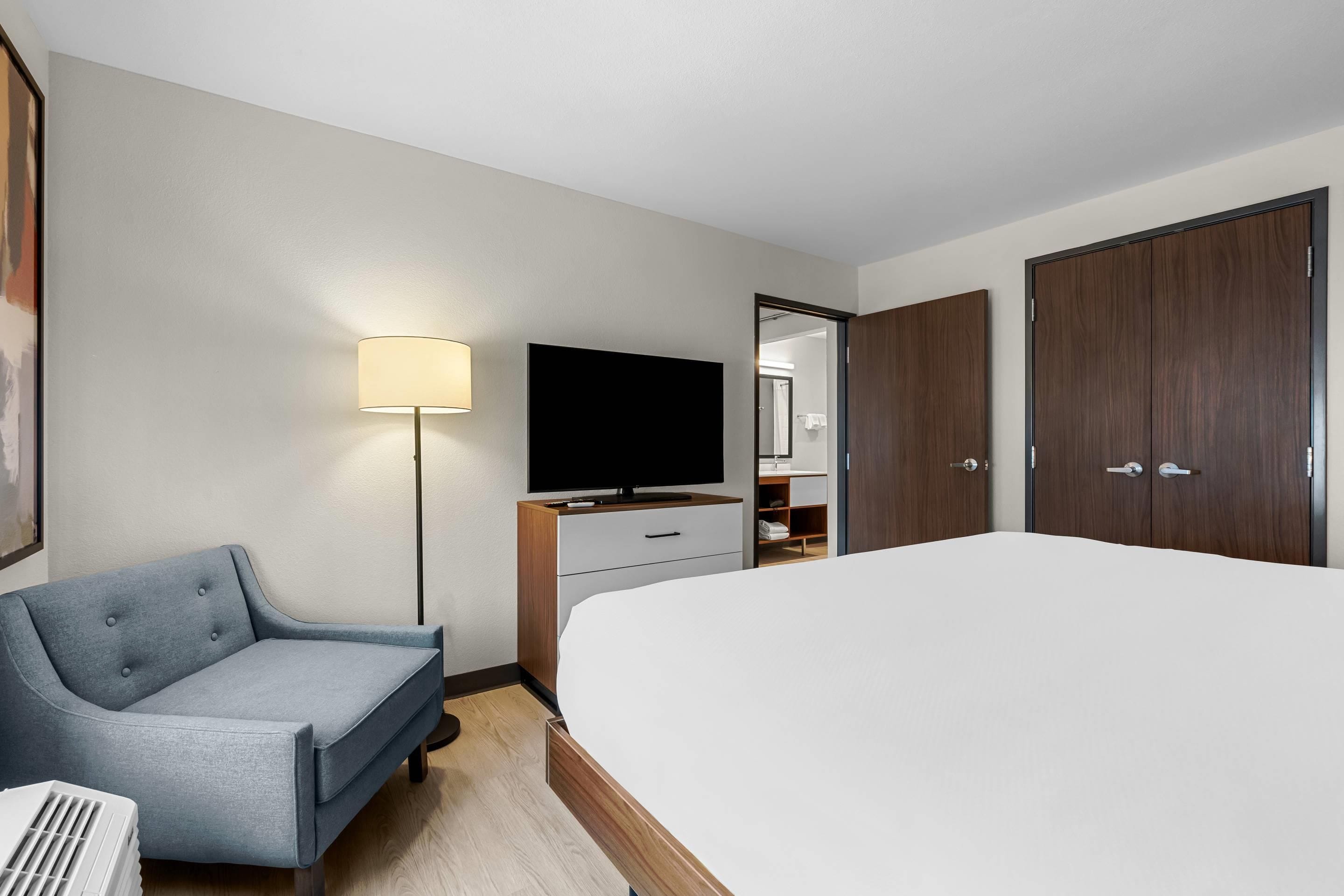 A hotel room with a bed, a desk with a flat-screen TV, a chair, a lamp, and a wardrobe.