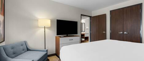 A hotel room with a bed, a desk with a flat-screen TV, a chair, a lamp, and a wardrobe.