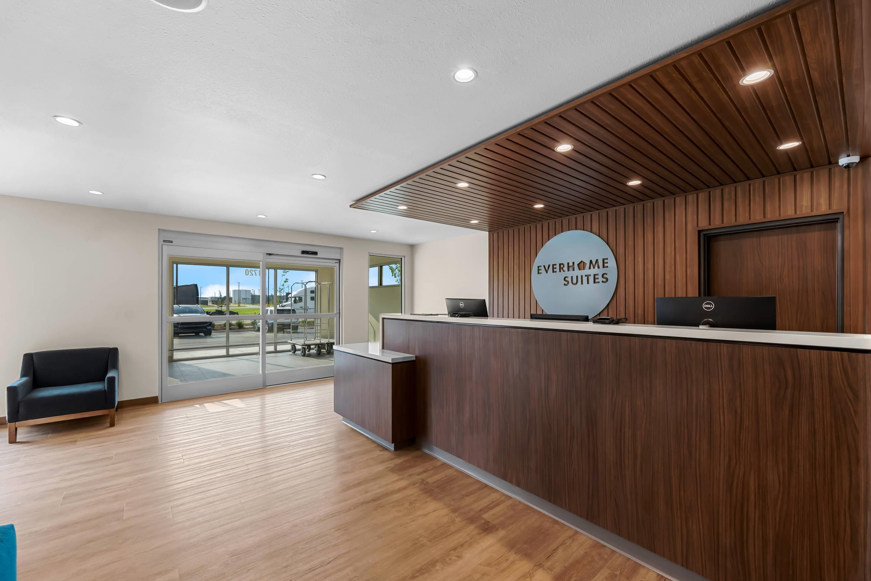 A reception desk with a sign for 'Everhome Suites' in a modern hotel lobby with wooden flooring and a glass door leading outside.