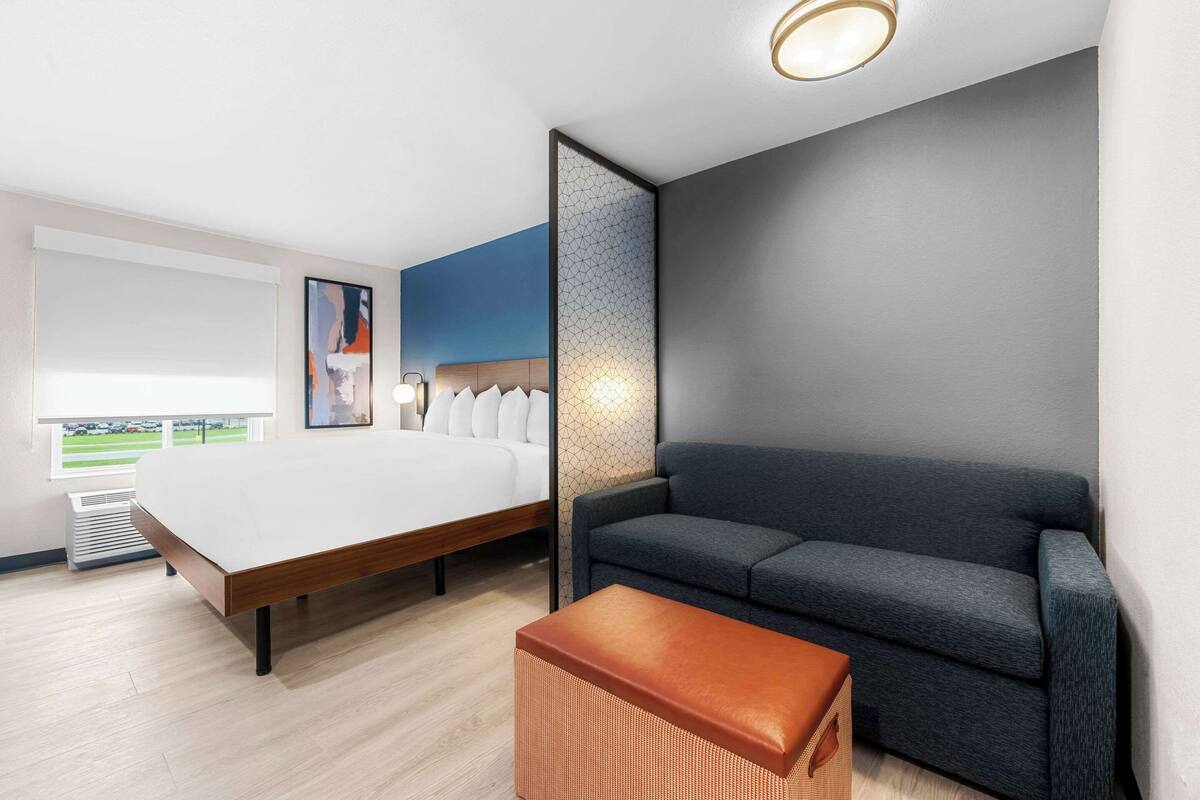 A modern hotel room with a large bed, a grey sofa, and a small ottoman.