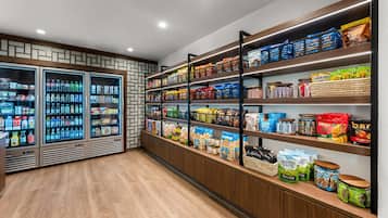 Interior de una tienda de comestibles con dispensadores refrigerados de bebidas, estantes llenos de snacks y productos envasados.