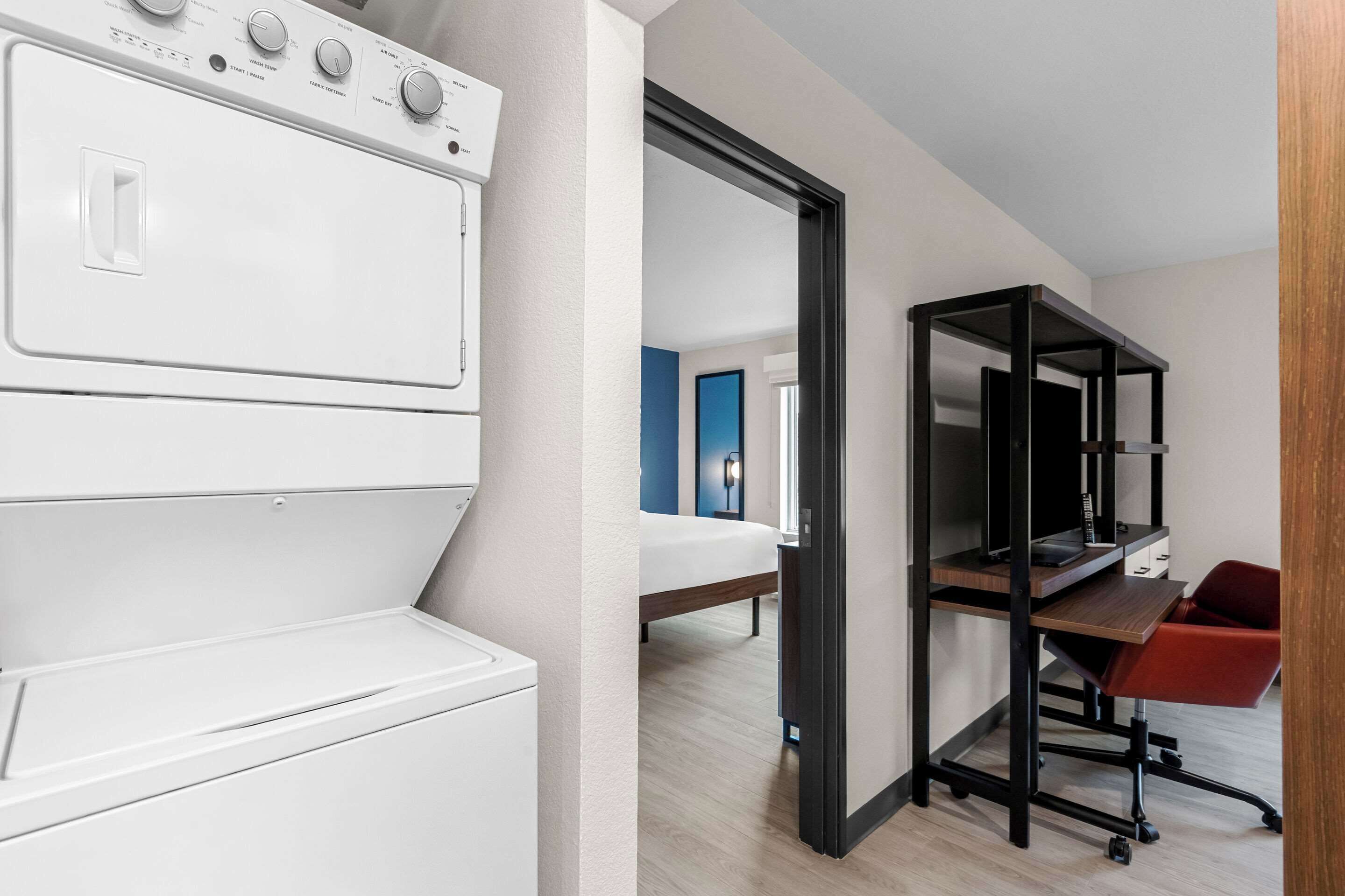 A modern hotel room with a built-in washer and dryer, a desk with a computer, and a red chair.