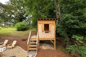 A wooden playhouse with a window and a small porch, surrounded by trees and a gravel path.