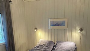 A single bed with a blue blanket and pillows, two wall-mounted lights, a framed picture on the wall, and a window with curtains. - Amazing place with boat, kayak, SUP and mountain view. Wifi and free parking.  (Nordland)