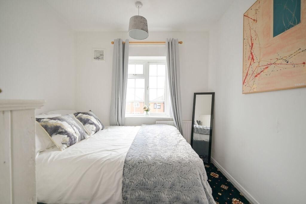 A bedroom with a bed, a window with curtains, a mirror, and a painting on the wall.
