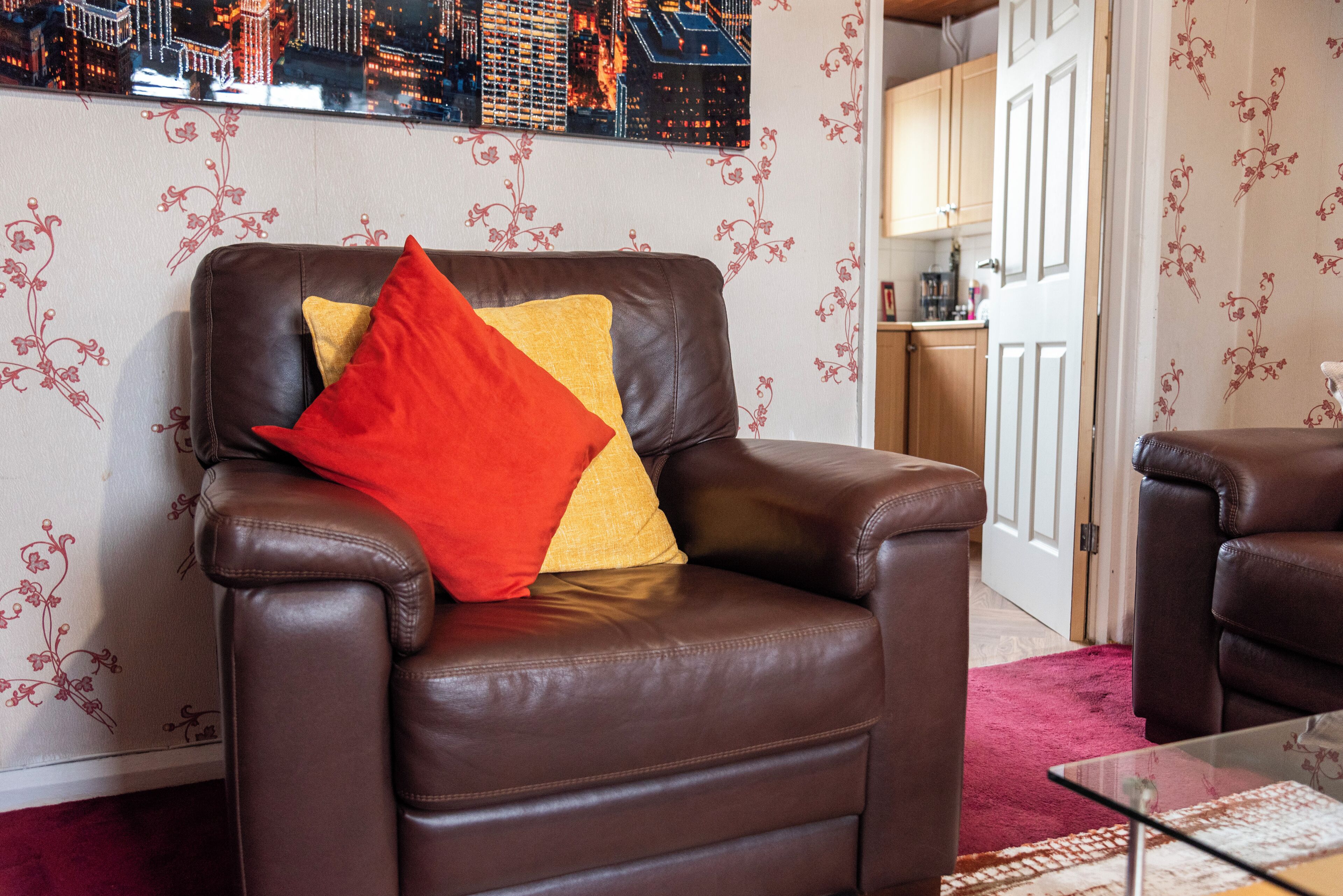 Un fauteuil en cuir marron avec un coussin rouge et jaune, dans une pièce dont le papier peint est à motifs floraux et où l’on peut voir un tableau représentant un paysage urbain accroché au mur.
