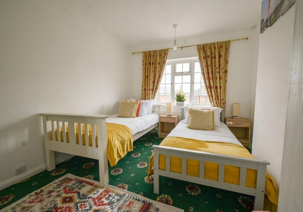 Une chambre avec deux lits simples, une petite table de chevet avec une lampe, une fenêtre avec des rideaux et un tapis à motifs sur le sol.