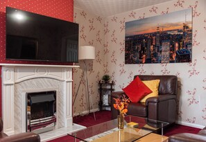 Een woonkamer met een open haard, een leren bank met rode en gele kussens, een flatscreen televisie boven de open haard en een stadslandschap schilderij aan de muur.