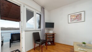 A room with a large window, a television on a wooden stand, a framed picture on the wall, and a small table with a plant.