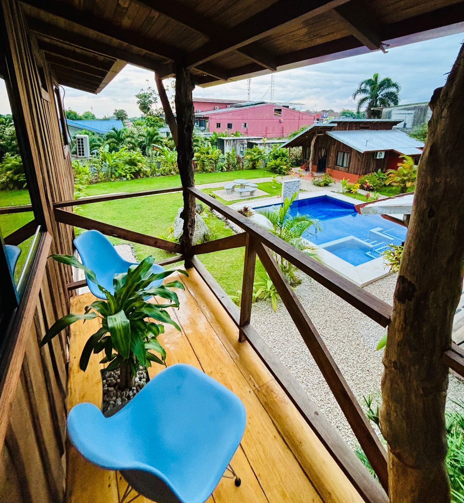 A wooden deck with two blue chairs, a small pool, and a view of a red building and greenery.