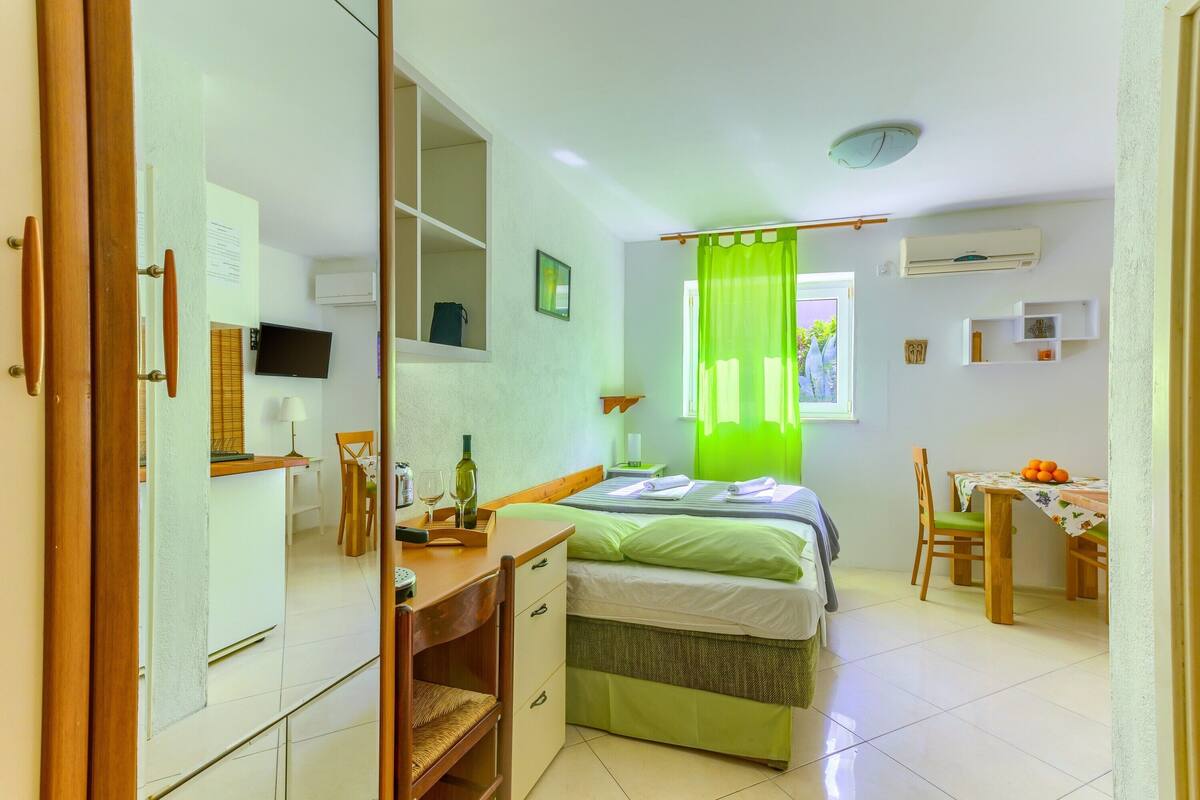 Un dormitorio con cama, escritorio, silla, mesa de comedor con sillas, televisor y ventana con cortina verde.