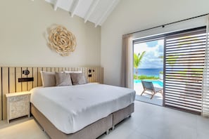 A modern bedroom with a large bed, a wooden headboard, a decorative wall piece, and a view of the ocean through a sliding door. - Luxurious oceanfront villa with private cinema and private pool! (Kralendijk)