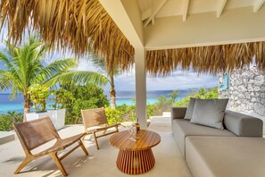 A modern outdoor seating area with a wooden table and chairs, a sofa, and a view of the ocean. - Luxurious oceanfront villa with private cinema and private pool! (Kralendijk)