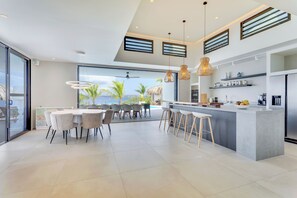 A modern kitchen with a central island, a dining area with a round table and chairs, and a view of the ocean through large windows. - Luxurious oceanfront villa with private cinema and private pool! (Kralendijk)