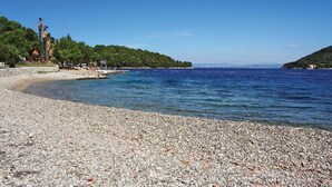 Une plage de galets bordée d’une mer d’un bleu limpide, une petite île couverte d’arbres et quelques bâtiments visibles au loin.