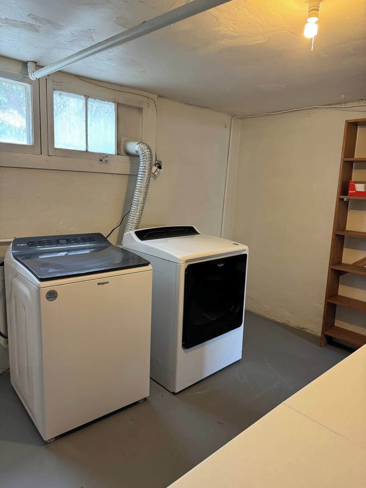 A laundry room with a front-loading washer and dryer, a wooden shelf, and a vent pipe.