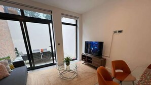Una sala de estar moderna con un sofá, una mesa de centro de vidrio y sillones naranja. Hay un televisor sobre un mueble de madera, y un ventanal con vistas al exterior.