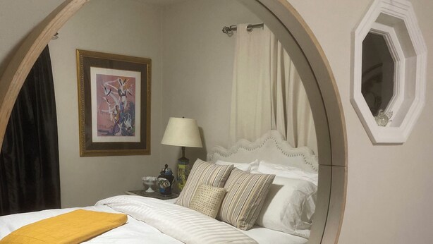 Ein runder Spiegel spiegelt ein Bett mit weißer Bettwäsche und einer gelben Tagesdecke, eine Nachttischlampe, ein gerahmtes Bild an der Wand und ein Fenster mit Vorhängen.