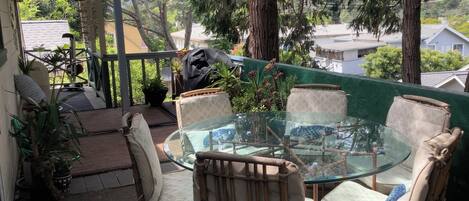 A covered patio with a glass table and chairs, surrounded by potted plants and a view of houses.