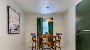 A dining area with a wooden table and chairs, a hanging light fixture, a window with green curtains, and a wall-mounted painting.