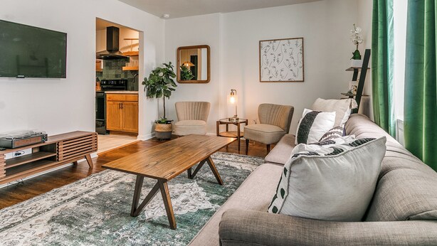 A modern living room with a wooden coffee table, two armchairs, a sofa, a ceiling fan, a TV, and a kitchen visible through an open door.