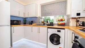 A compact kitchen with white cabinets, a built-in washing machine, a microwave, and a window with a view of greenery. - Spacious 2BR Flat @ Summertown | Sleeps 7 | 5 Star (Oxford)