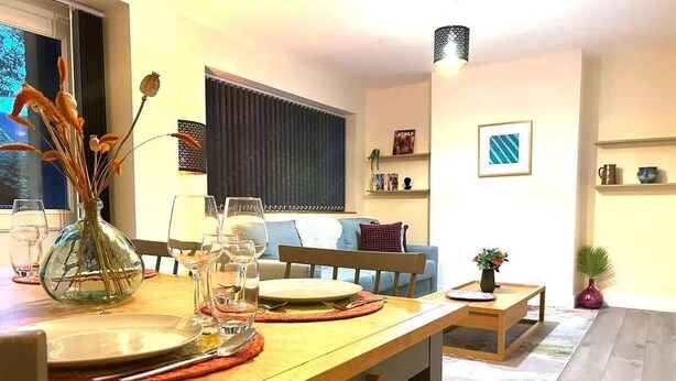 A dining area with a wooden table set for two, a vase with dried flowers, and a window with curtains. - Spacious 2BR Flat @ Summertown | Sleeps 7 | 5 Star (Oxford)