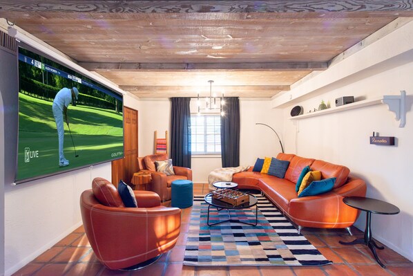 Un salon moderne doté d’un grand téléviseur diffusant un match de golf, d’un confortable canapé orange, d’un pouf bleu et d’un tapis à motifs.