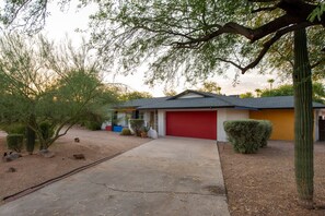 Une maison d’habitation avec une porte de garage rouge, une allée en béton et un petit jardin agrémenté de diverses plantes et d’un cactus.