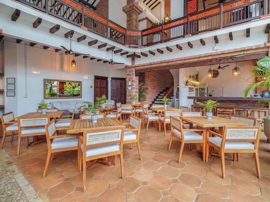 Un amplio patio al aire libre con mesas y sillas de madera, un muro de ladrillo y una escalera que lleva a un nivel superior.