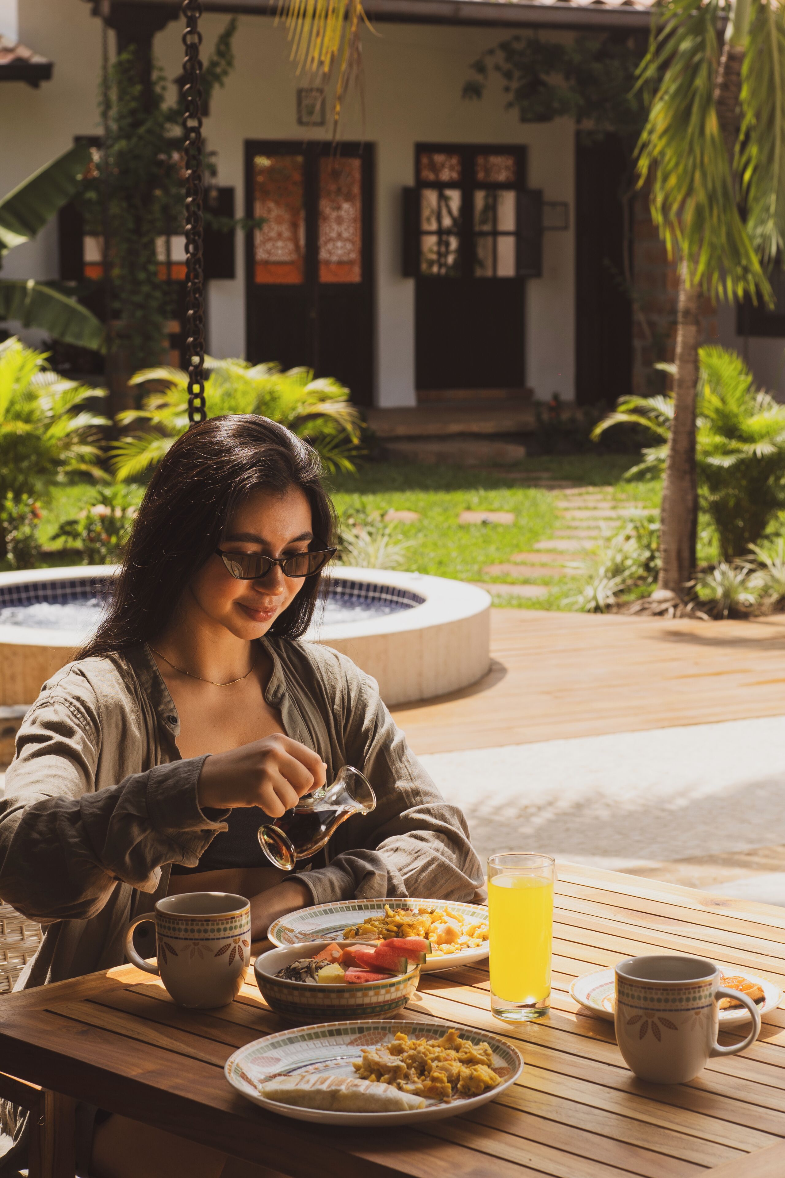 A person sitting at an outdoor table with a meal, a teapot, and a glass of juice.