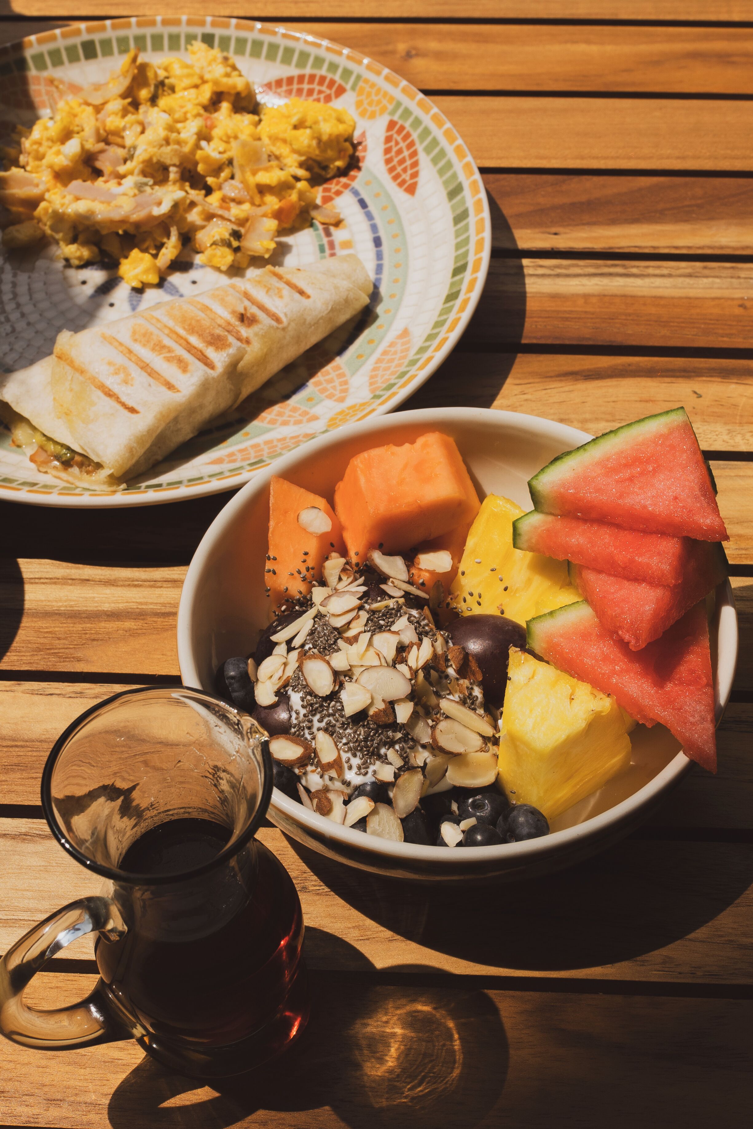 A meal with a plate of scrambled eggs, a wrap, and a bowl of fruit salad.