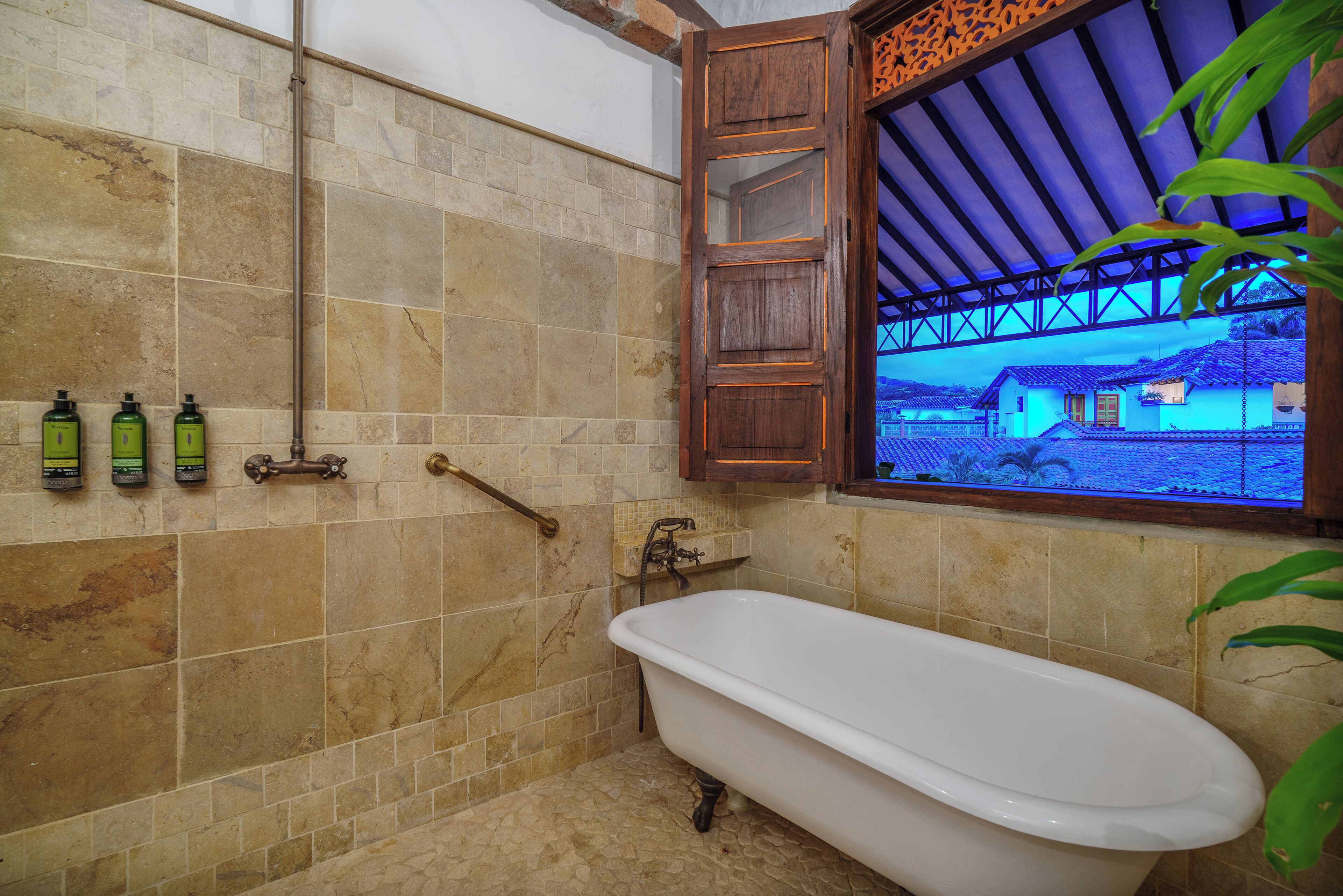 Een badkamer met een bad, betegelde wanden, een raam met uitzicht op een huis en toiletartikelen aan de muur.