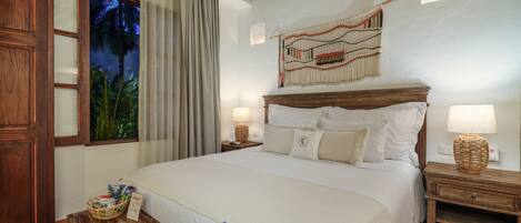 A neatly arranged hotel room with a large bed, two bedside tables with lamps, a wooden bench, a decorative wall hanging, and a window with curtains.