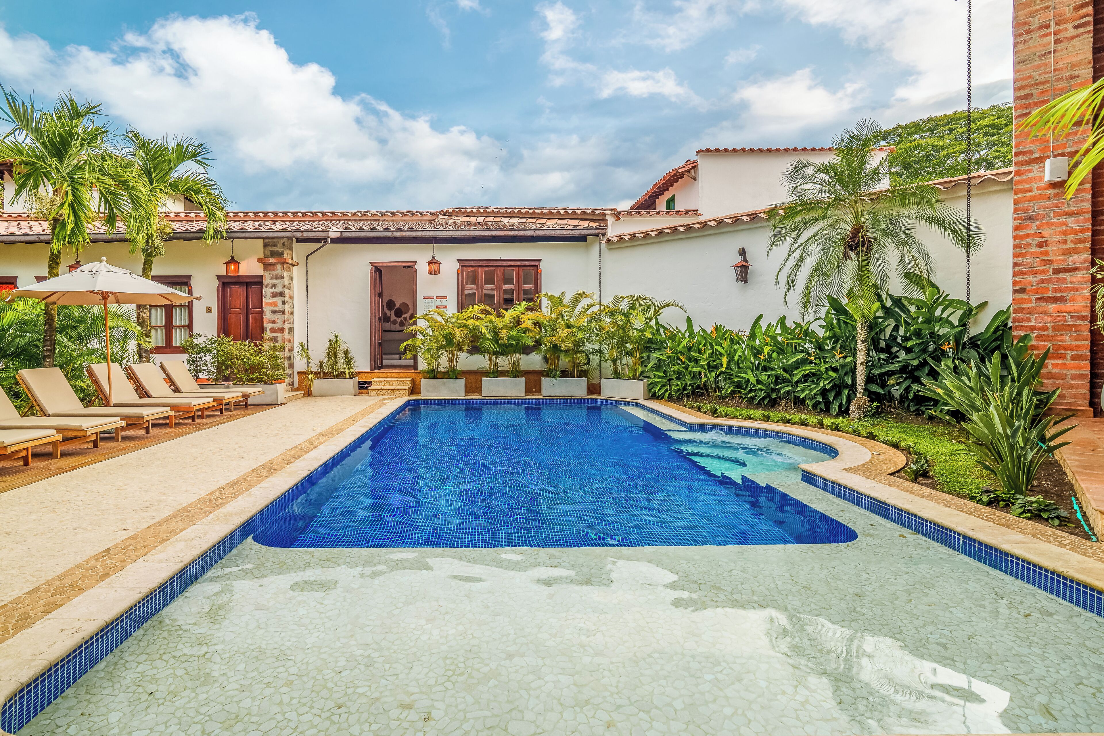 A swimming pool with lounge chairs and palm trees, surrounded by a building with a tiled roof and a door.