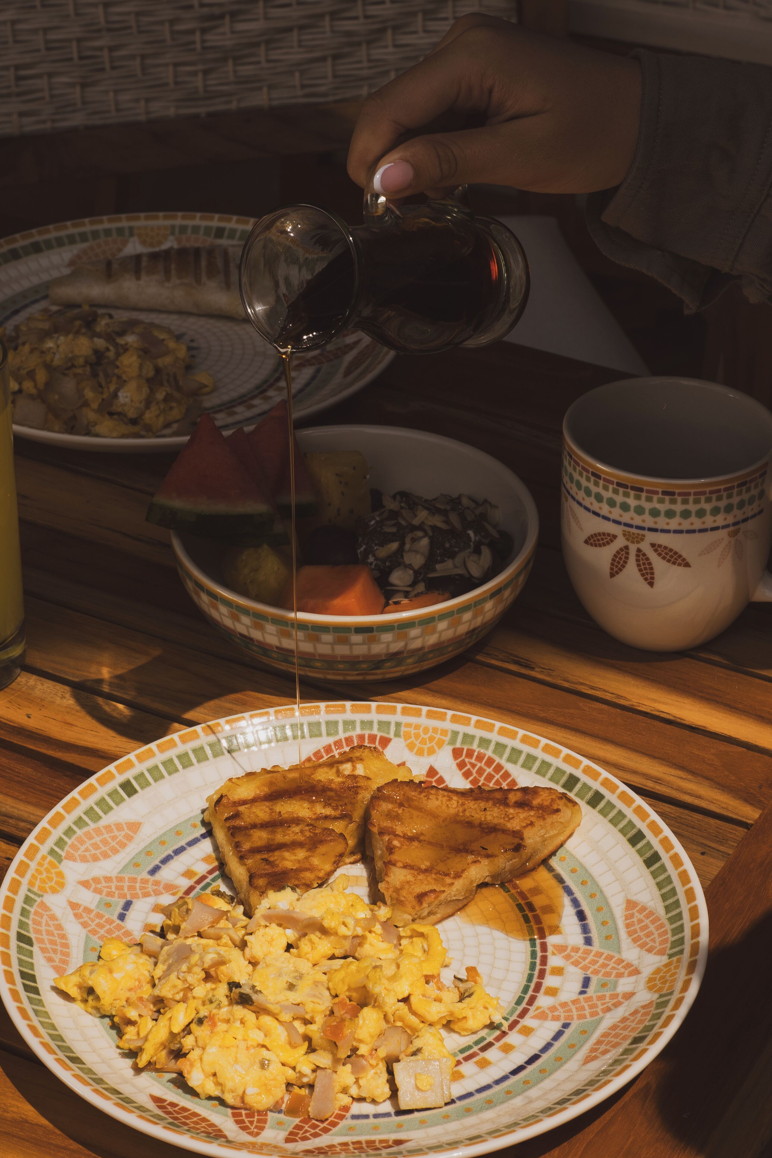 Une assiette de petit-déjeuner avec des toasts et des œufs brouillés, un bol de fruits et une tasse de café, le tout posé sur une table en bois.