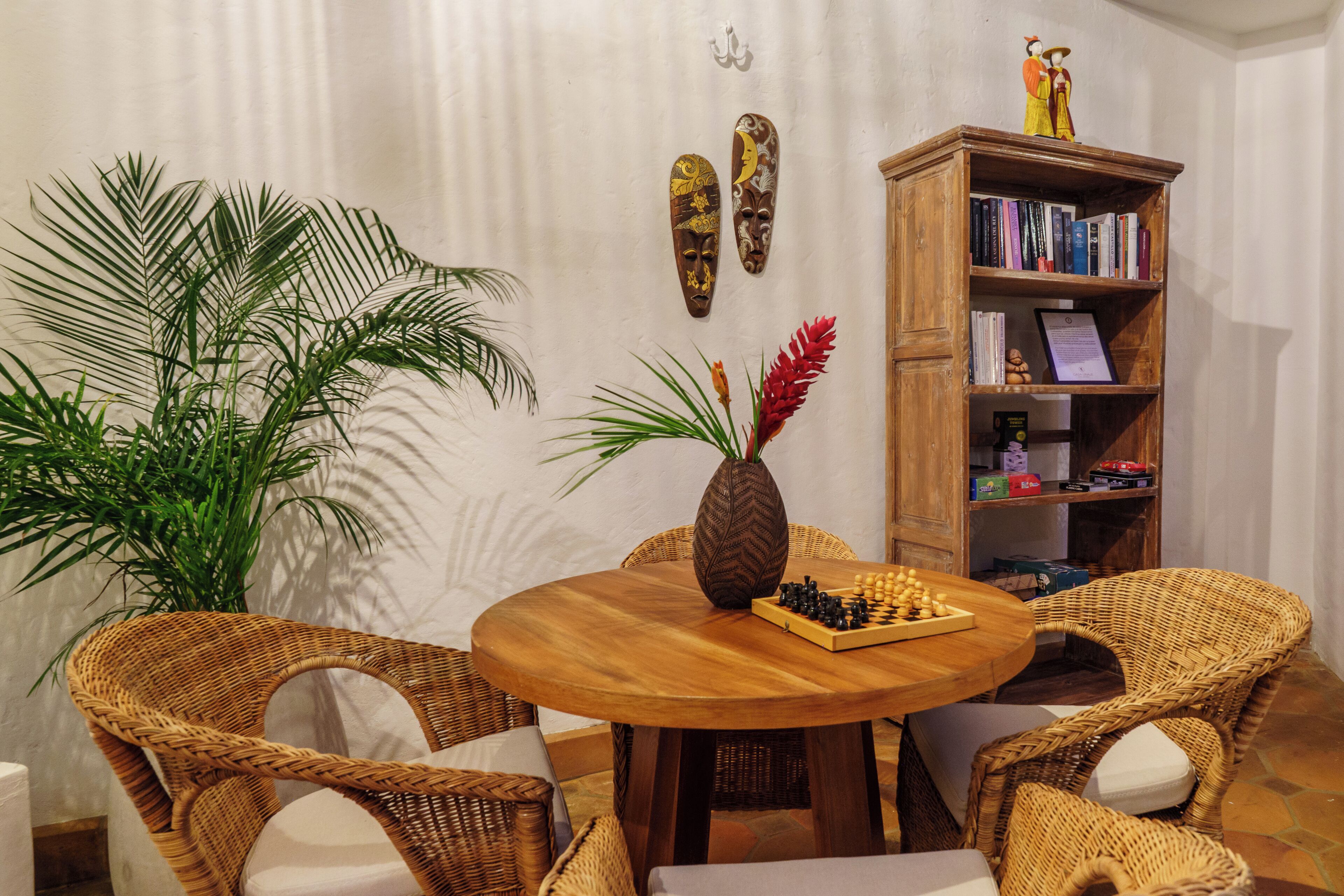 Een ronde houten tafel met een schaakbord, omringd door rieten stoelen, een grote potplant en een houten boekenkast met boeken en decoratieve objecten.