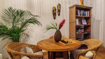 Een ronde houten tafel met een schaakbord, omringd door rieten stoelen, een grote potplant en een houten boekenkast met boeken en decoratieve objecten.
