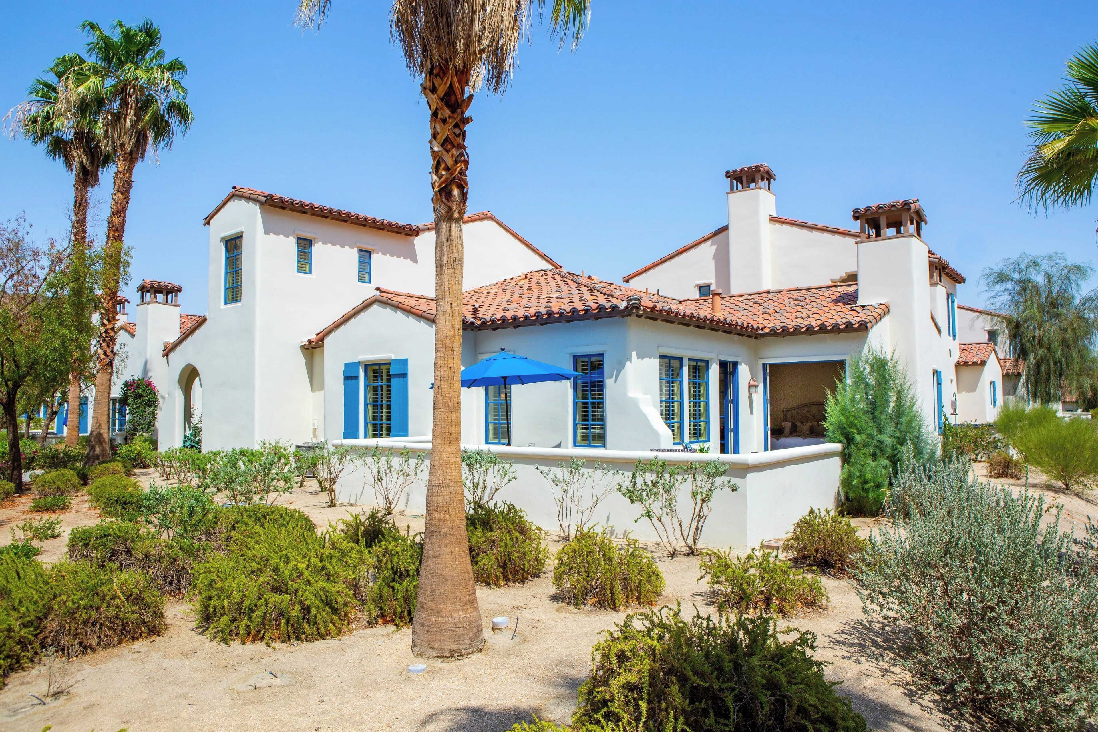 A white building with blue shutters and a tiled roof, surrounded by various plants and palm trees.