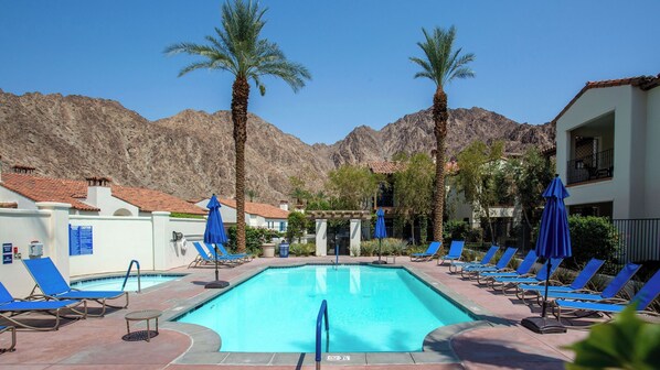A swimming pool with lounge chairs and umbrellas, surrounded by palm trees and mountains.
