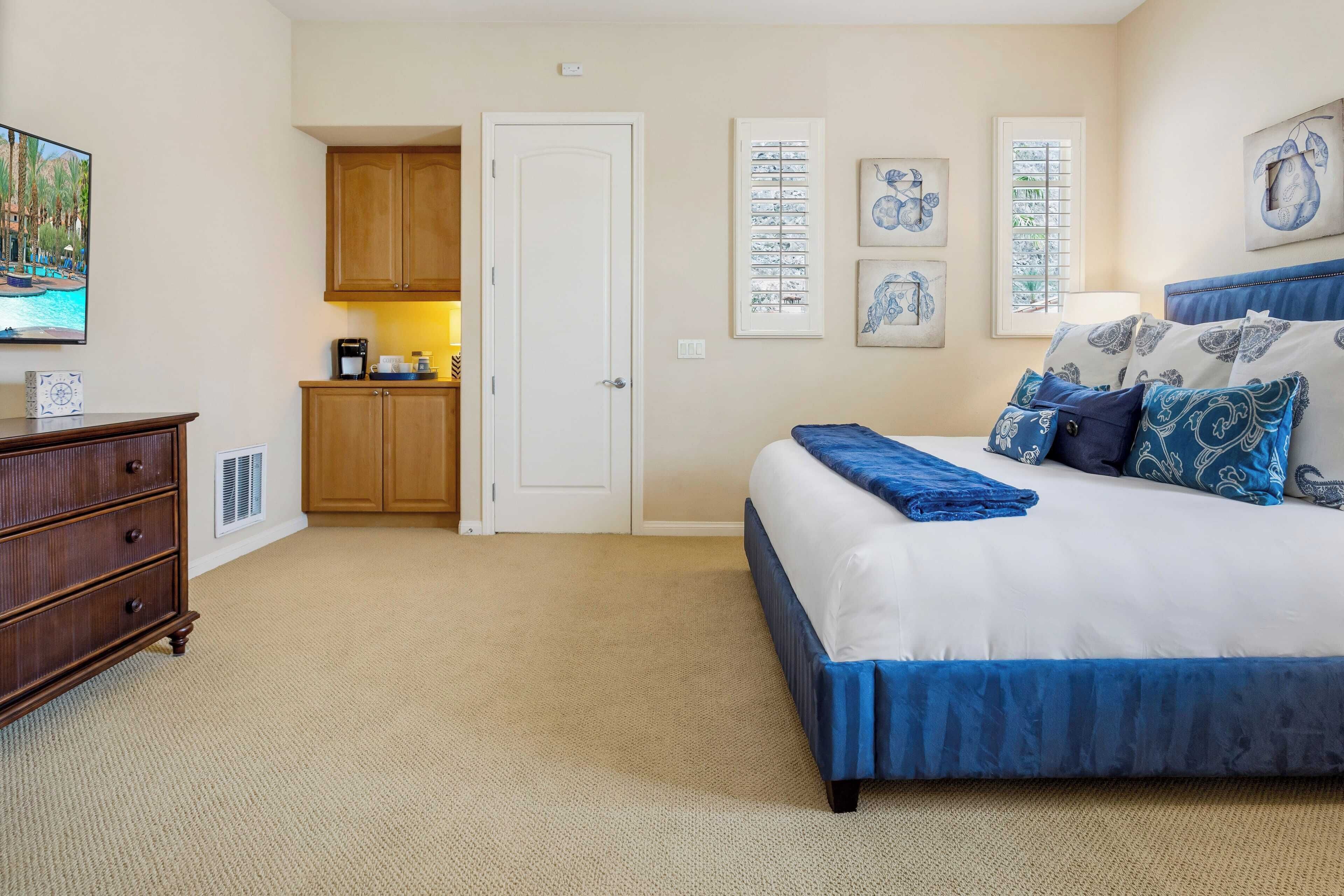 A bedroom with a large bed, a dresser, a small kitchenette, and artwork on the walls.
