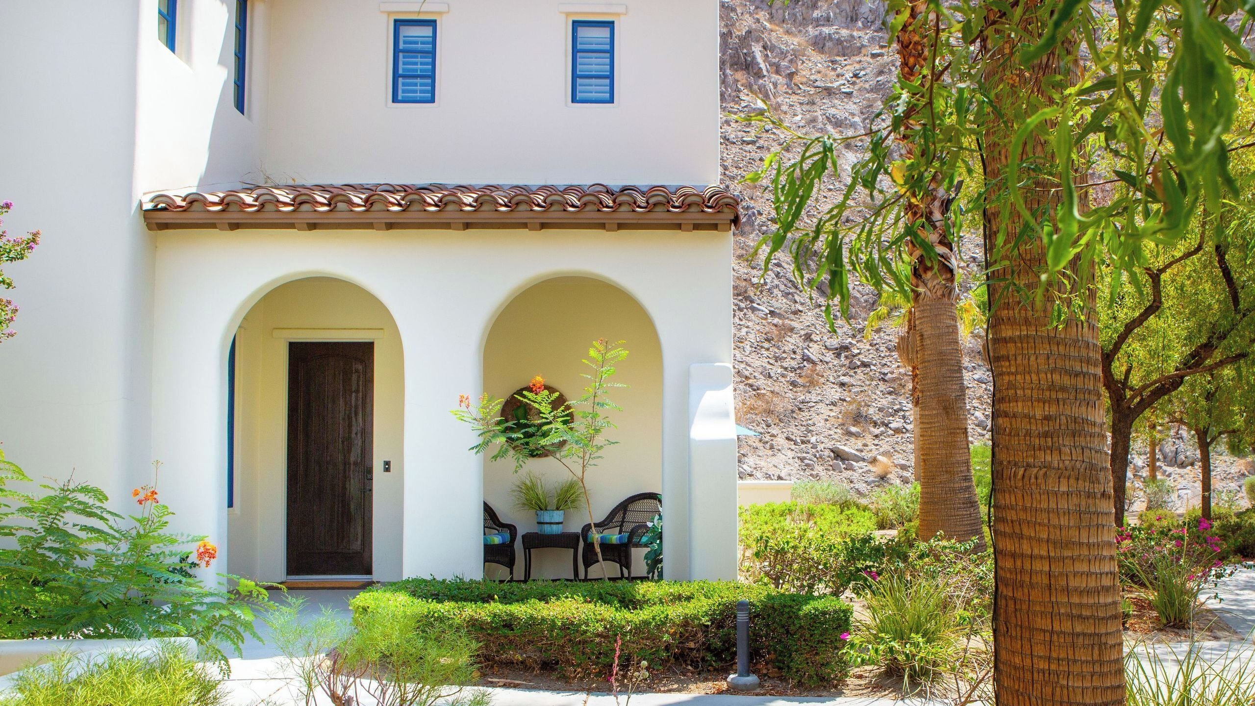 A building with a white facade, two blue windows, a brown door, and a small porch with a bench and potted plants.