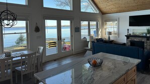 Amplia sala de estar con ventanas grandes con vistas a un lago, chimenea de piedra y cocina moderna con una isla de mármol.