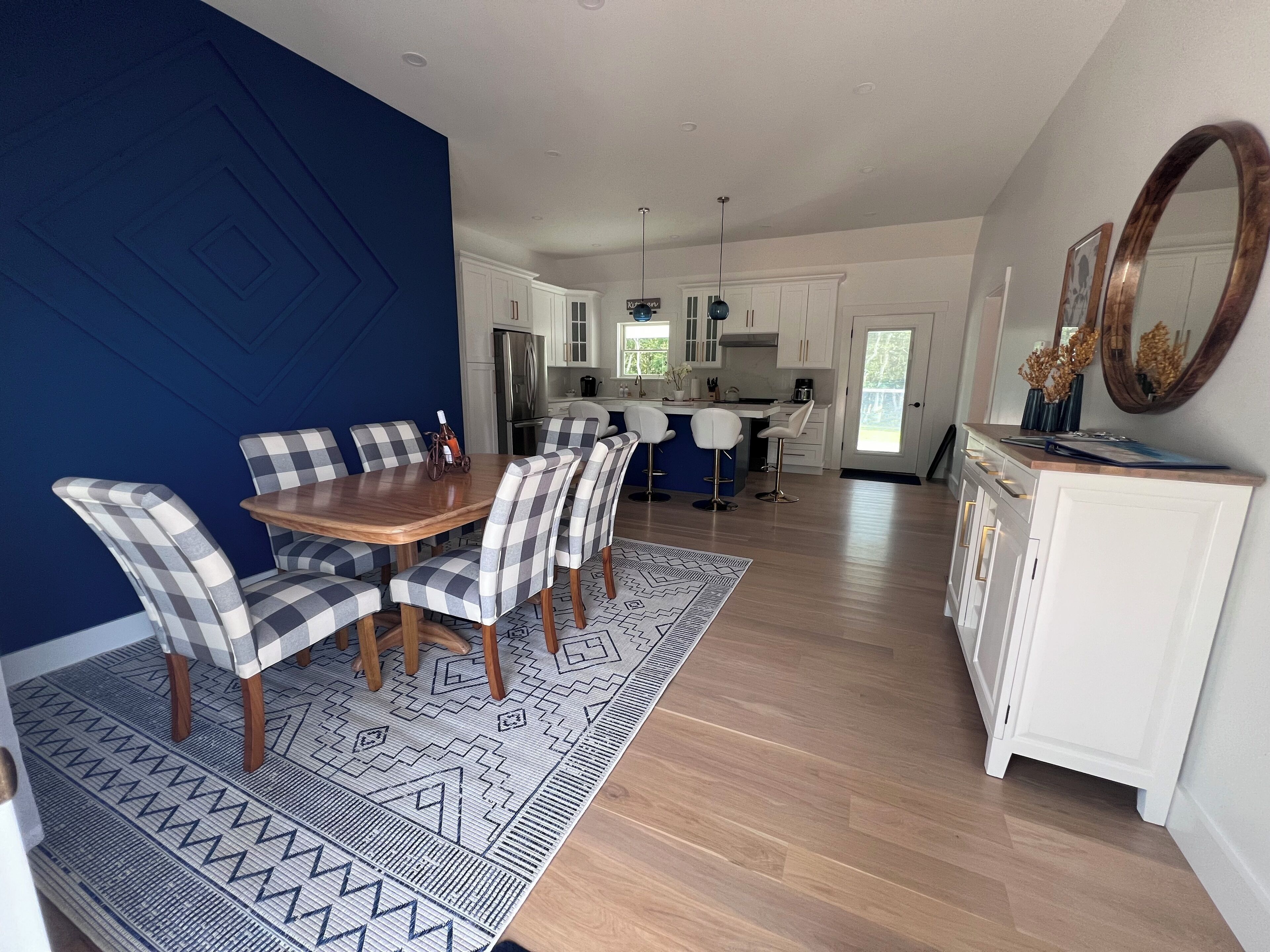 Un coin repas avec une table en bois et des chaises à carreaux, une cuisine avec des meubles blancs et un grand miroir au mur.