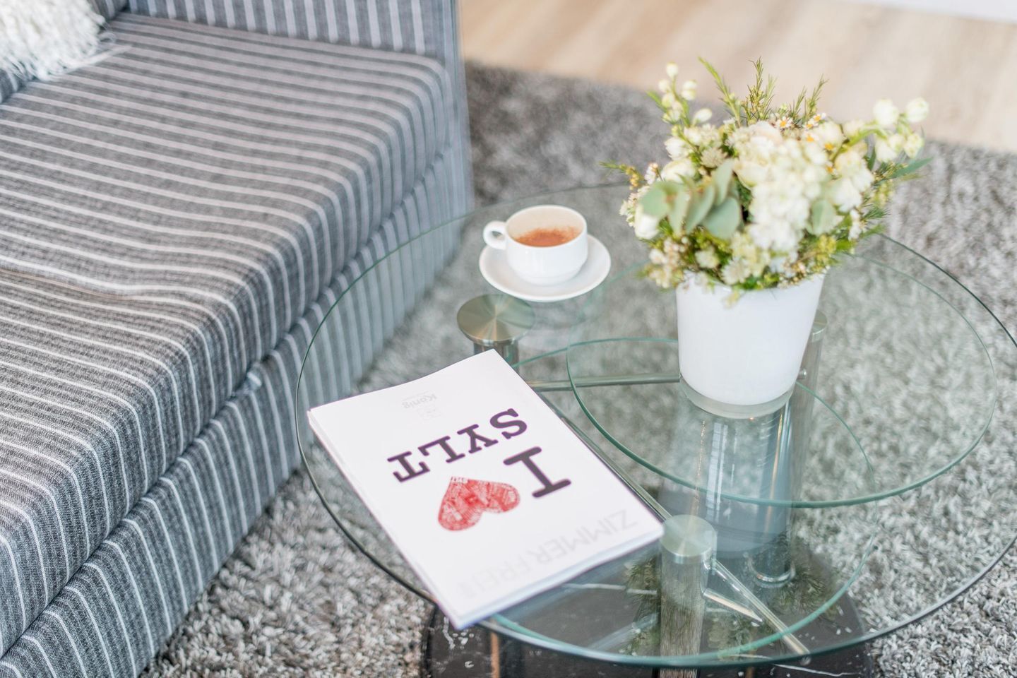 A modern living room with a striped grey sofa, a glass coffee table with a book titled "STYLE I", a white flower vase, and a cup of coffee.