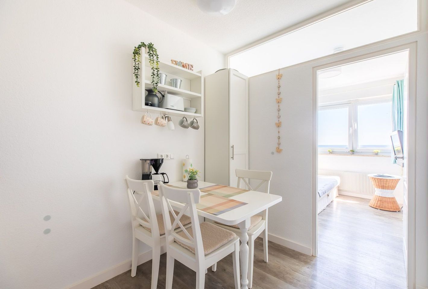 A bright, well-lit room with a white table and chairs, a coffee maker, and a shelf with kitchen items.