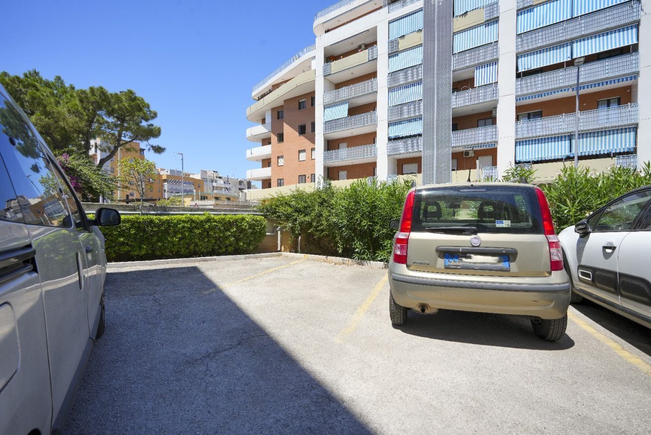 Un parking avec une voiture beige, une voiture blanche et un immeuble doté de nombreux balcons.