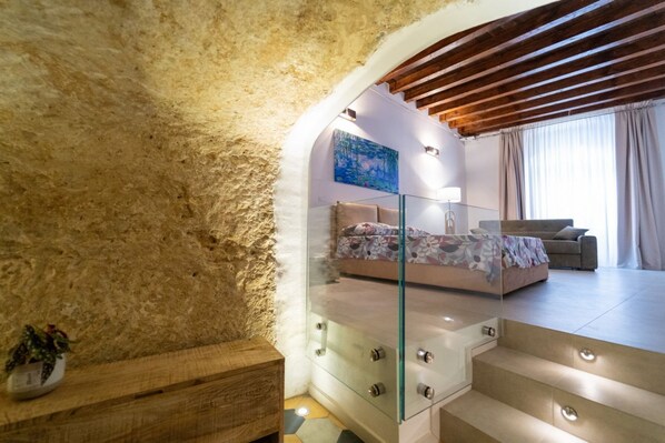 A bedroom with a stone wall, a glass staircase, a bed with a floral bedspread, a sofa, and a painting on the wall.